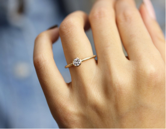 Minimalist solitaire engagement ring with a 0.25 carat lab grown diamond in a yellow gold band, worn on a hand against a denim background.
