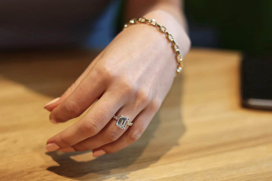 Hand wearing an emerald-cut lab grown diamond ring with a halo setting, styled casually on a wooden table.