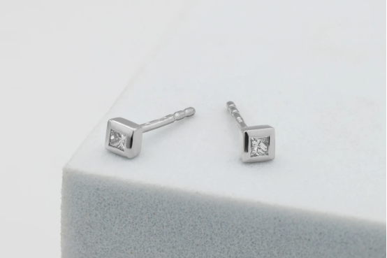 Princess-cut lab grown diamond stud earrings in a bezel setting, displayed on a light grey foam stand.