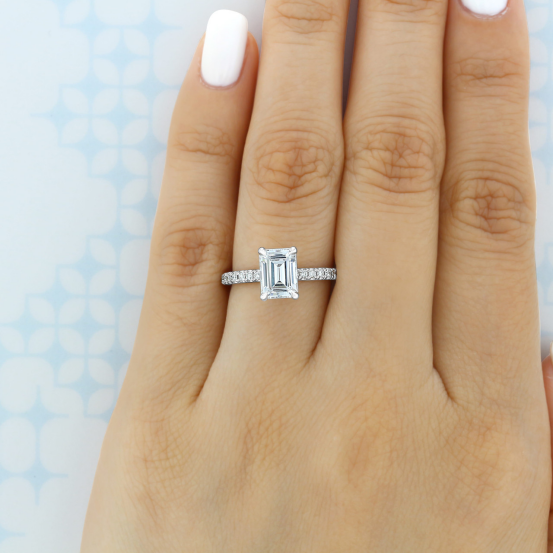 Hand with white nail polish wearing an emerald-cut lab grown diamond engagement ring with a pavé-set band, against a light blue patterned background.